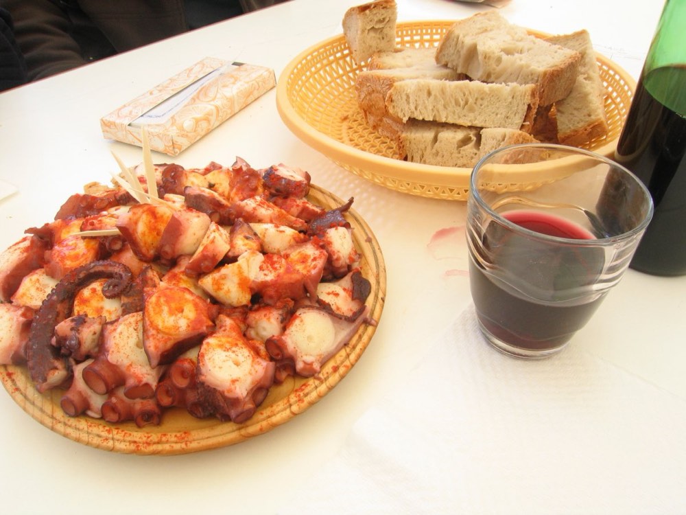 Polbo á feira served on a wooden plate with paprika, next to a basket of bread and a glass of red wine.